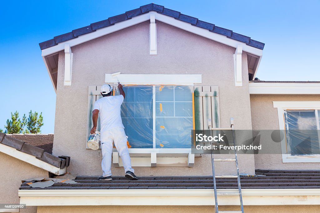 Un pintor profesional vestido de blanco trabaja en la fachada de una vivienda de dos niveles, utilizando un rodillo para aplicar pintura blanca sobre el marco de una ventana protegida con plástico, como parte de un servicio pintura de casas