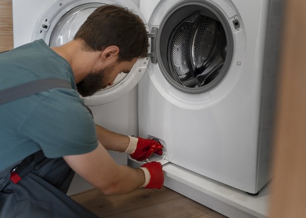 Technician in teal shirt and red gloves kneels beside an open-front washing machine, performing maintenance at the base panel.