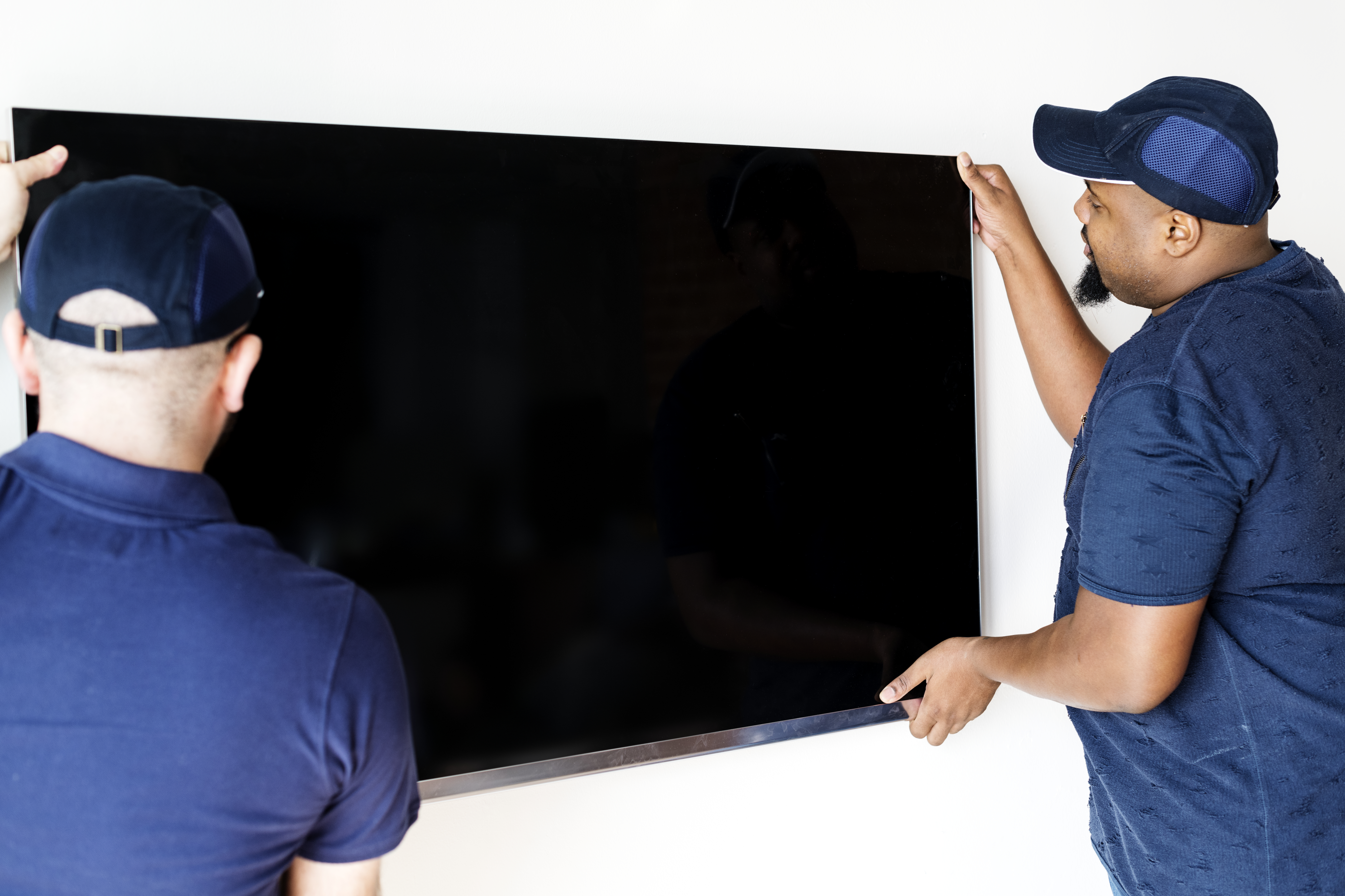 Dos técnicos profesionales con gorra y camisetas azules sostienen con cuidado una gran pantalla plana durante el proceso de instalación de televisión en una pared blanca.