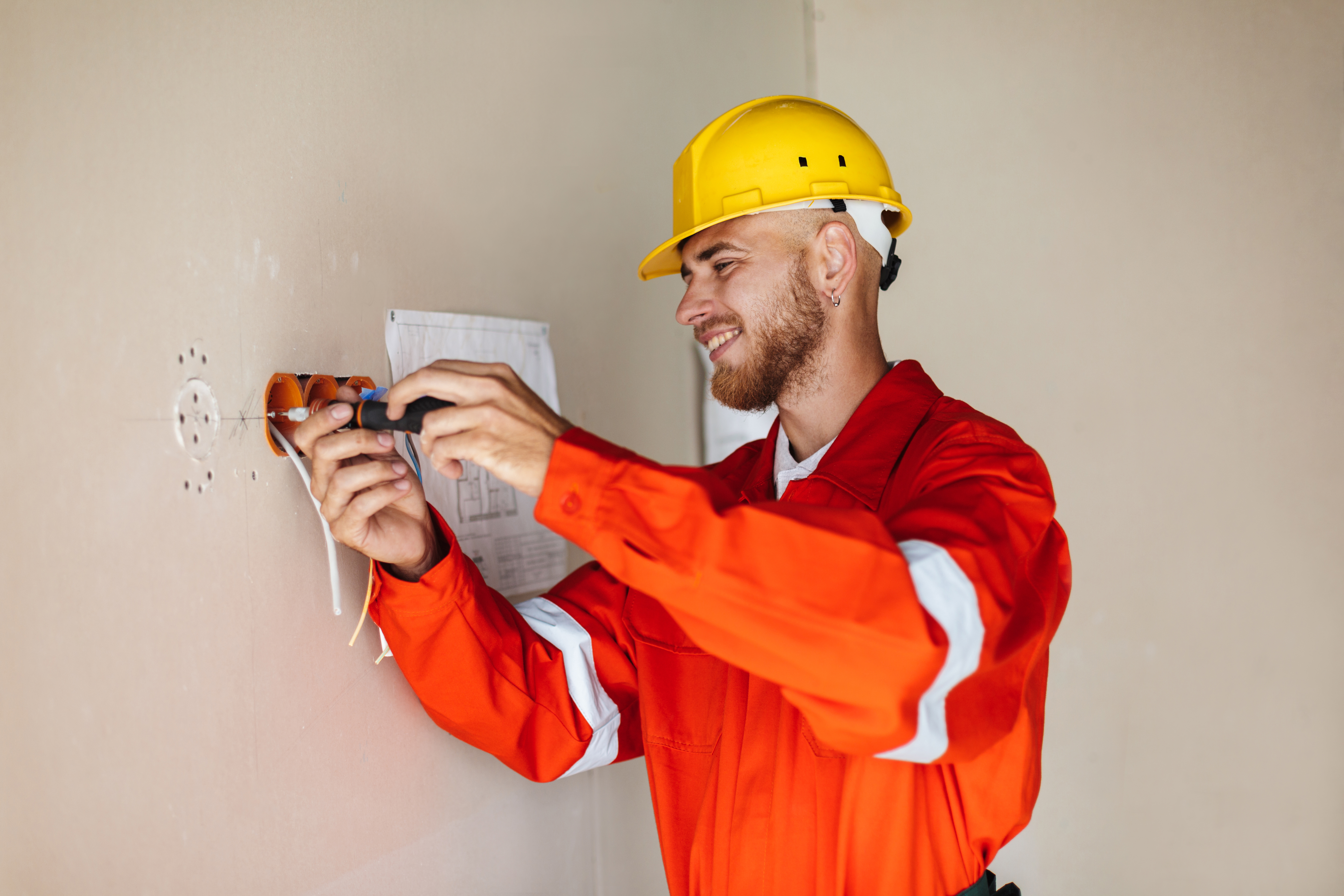 Un electricista sonriente con overol naranja y casco amarillo trabaja en la pared instalando una base de enchufe como parte de una instalación eléctrica en casa Un electricista sonriente con overol naranja y casco amarillo trabaja en la pared instalando una base de enchufe como parte de una instalación eléctrica en casa