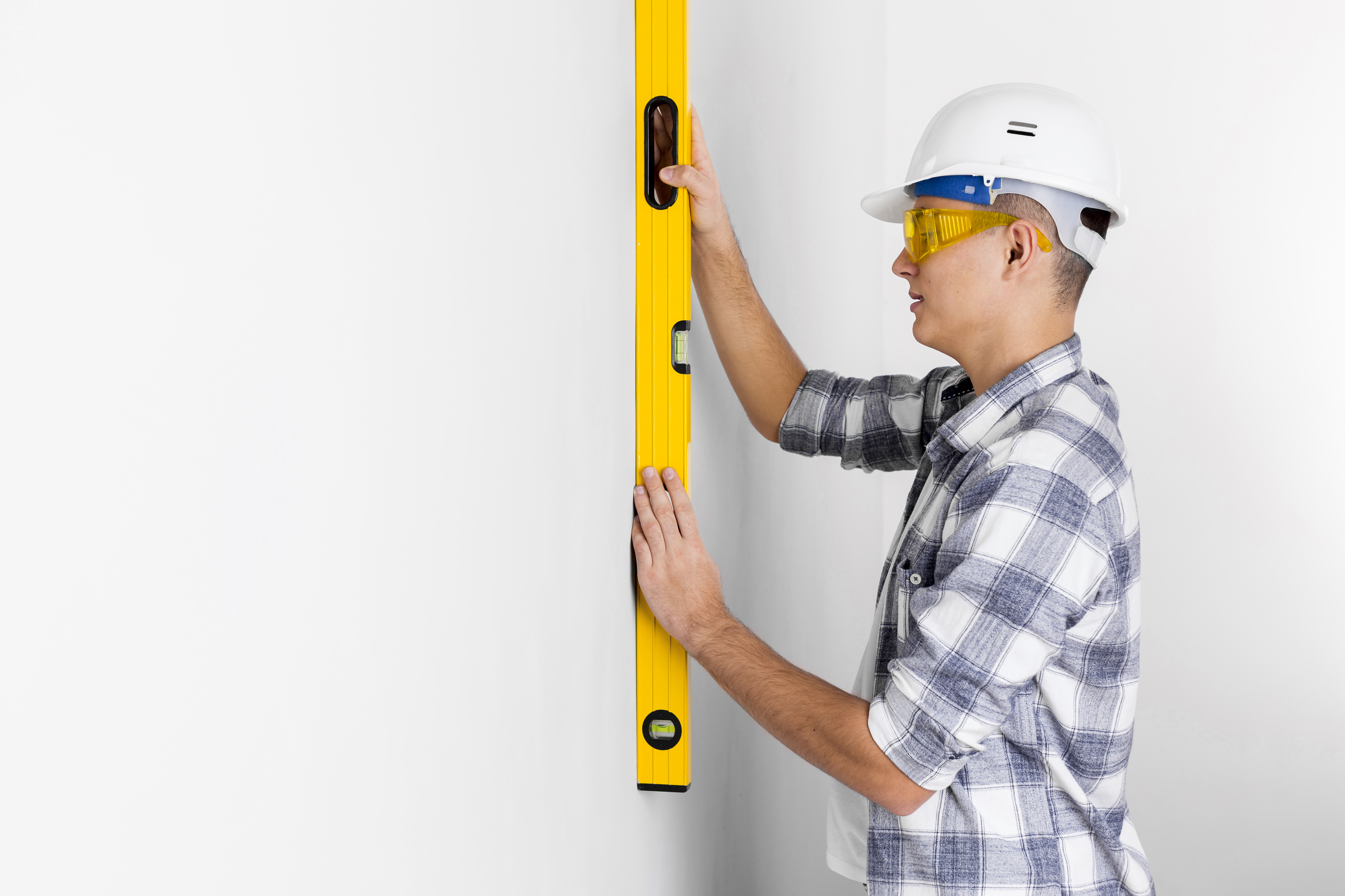 Profesional con casco blanco y gafas de seguridad utiliza un nivel de burbuja amarillo sobre una pared blanca para asegurar la alineación antes de calcular metros cuadrados en una remodelación