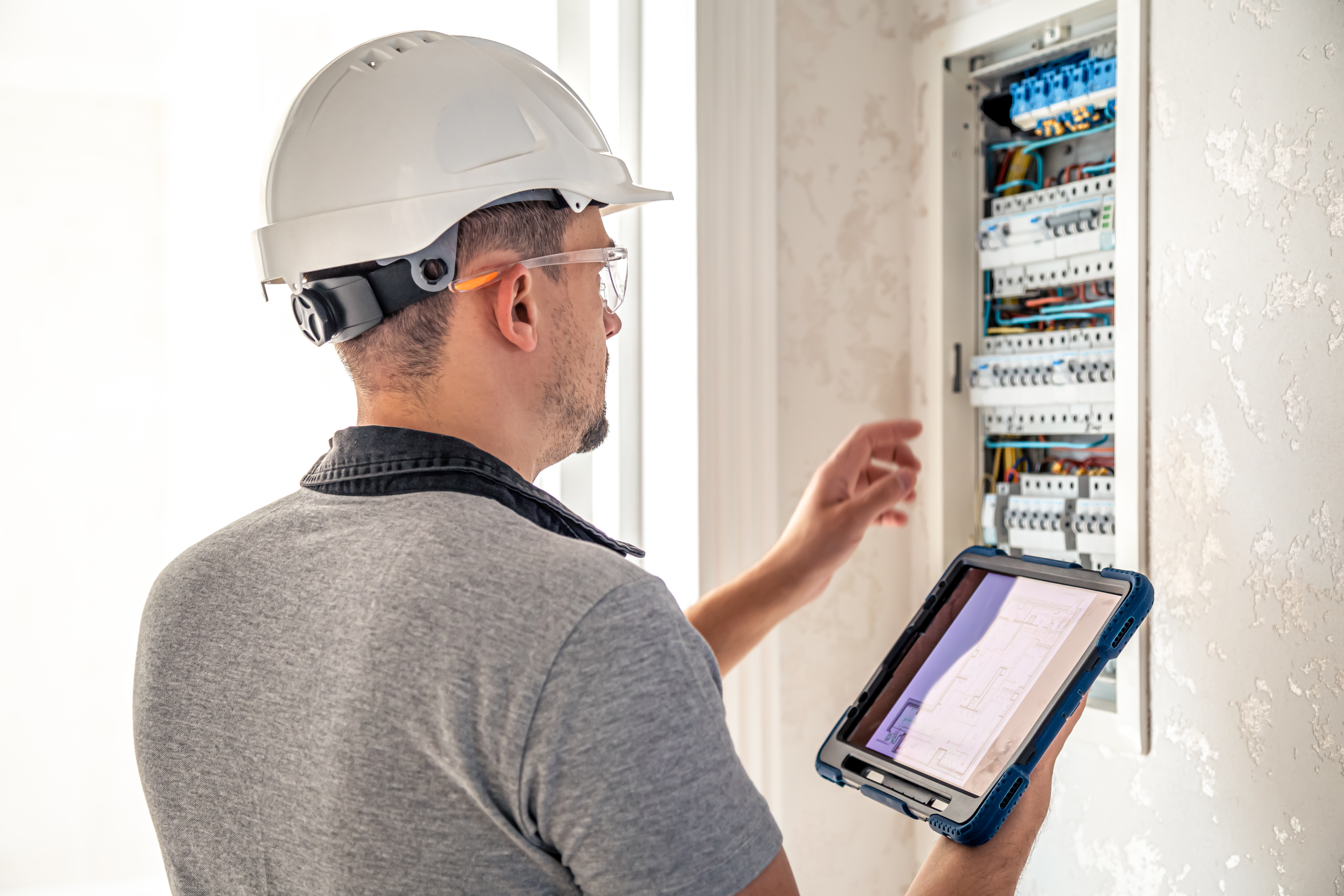 Un electricista con casco blanco y gafas de seguridad examina un cuadro eléctrico residencial mientras sostiene una tablet con un plano para una instalación eléctrica en casa Un electricista con casco blanco y gafas de seguridad examina un cuadro eléctrico residencial mientras sostiene una tablet con un plano para una instalación eléctrica en casa
