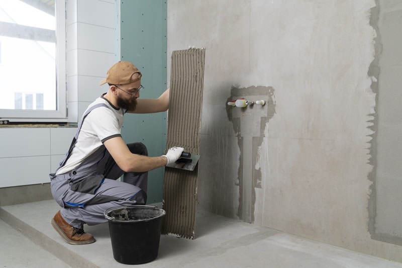 Hombre instalando azulejos grises para remodelar baños pequeños Hombre instalando azulejos grandes de color gris en el muro de un baño moderno, ejemplificando el proceso de remodelar baños pequeños con acabados elegantes
