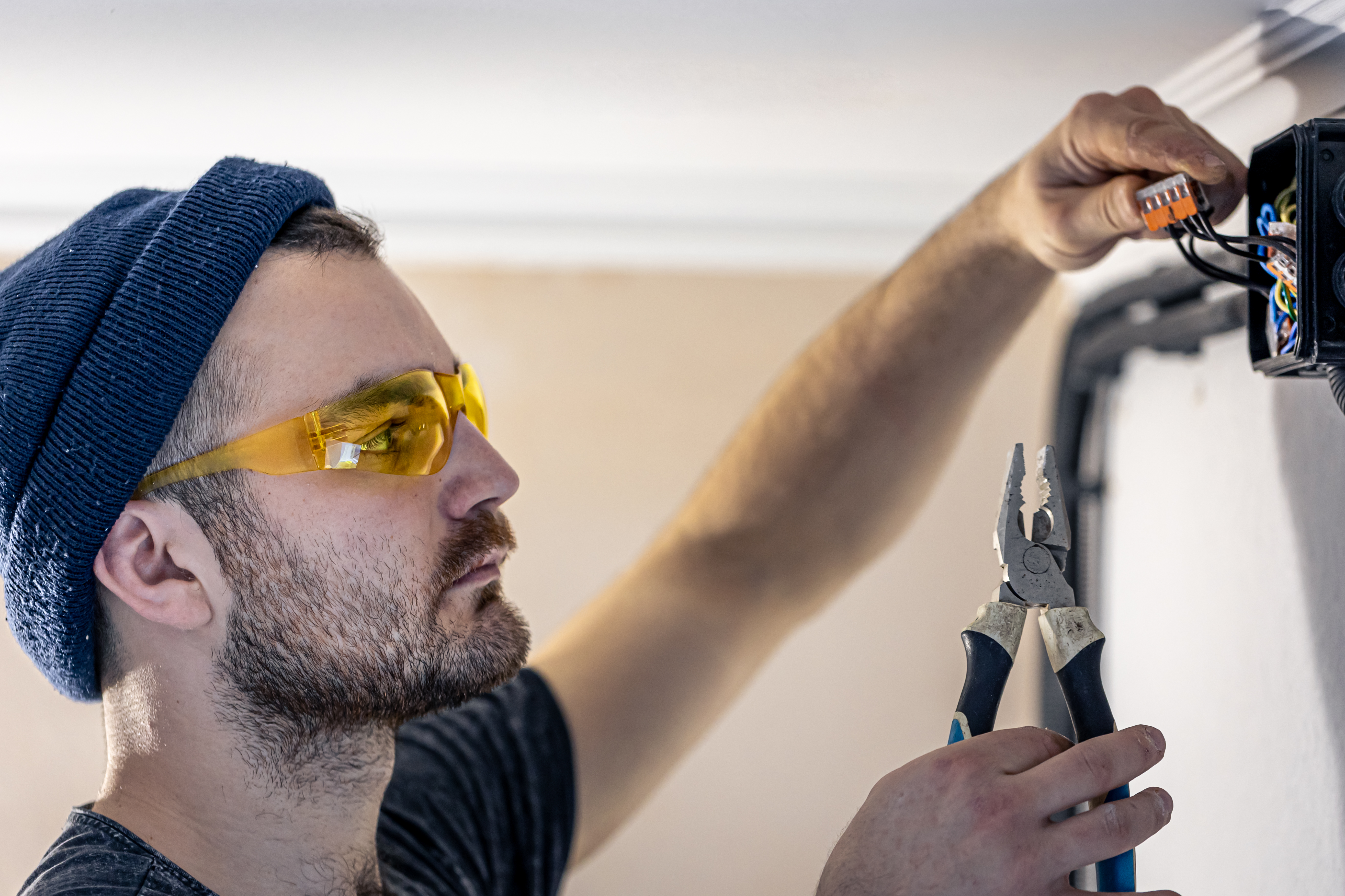 Un técnico eléctrico de emergencia con gafas de protección y herramientas realizando un servicio express de reparación de cables en una caja de conexiones de pared.